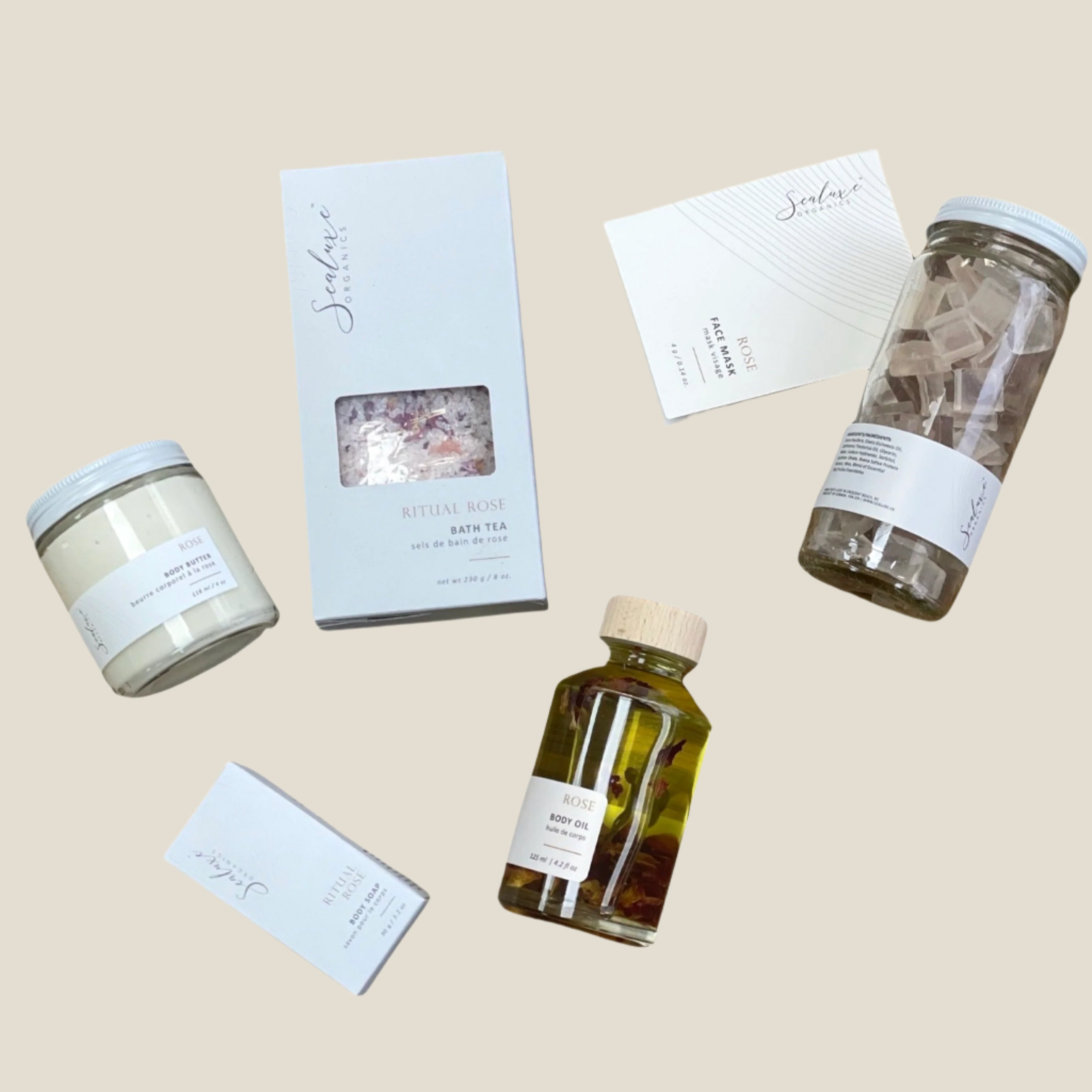 A collection of rose-scented skincare products including a rose mask, body butter, body oil, bath salts, and soap bars, packaged in various containers such as jars and boxes.