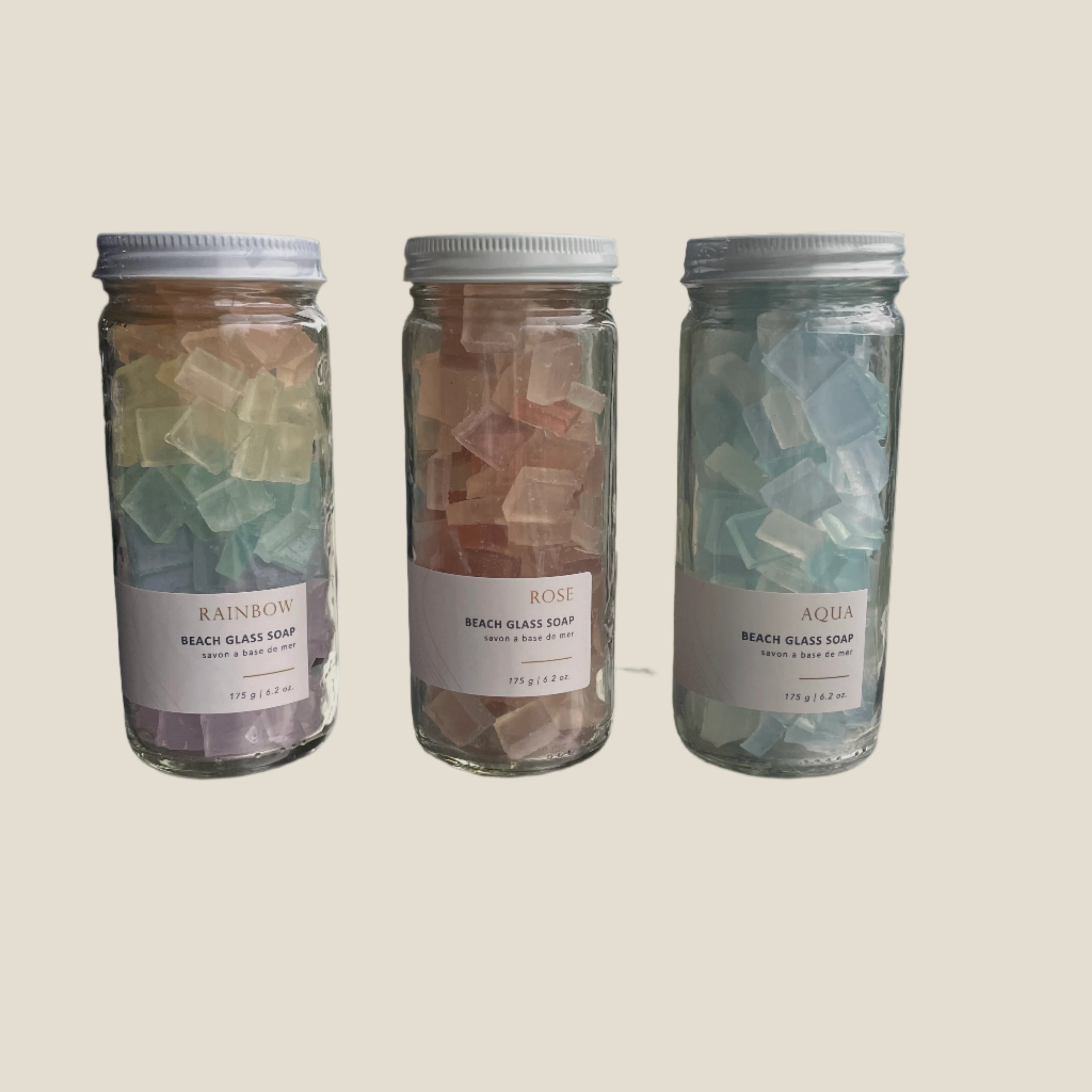 Three jars of beach glass soap labeled 'Rainbow', 'Rose', and 'Aqua' on a surface near a window.