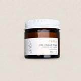 A small amber glass jar candle with a white screw-top lid and a white label reading TREES FIR + BLACK PINE coconut soy candle, centered on a warm beige background.
