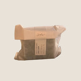 A wrapped rectangular body soap bar sits on a windowsill with a blurred outdoor background. The soap is covered in a light beige paper sleeve with a white paper band label that reads 'Serenade Soaps' and 'Activated Charcoal'.