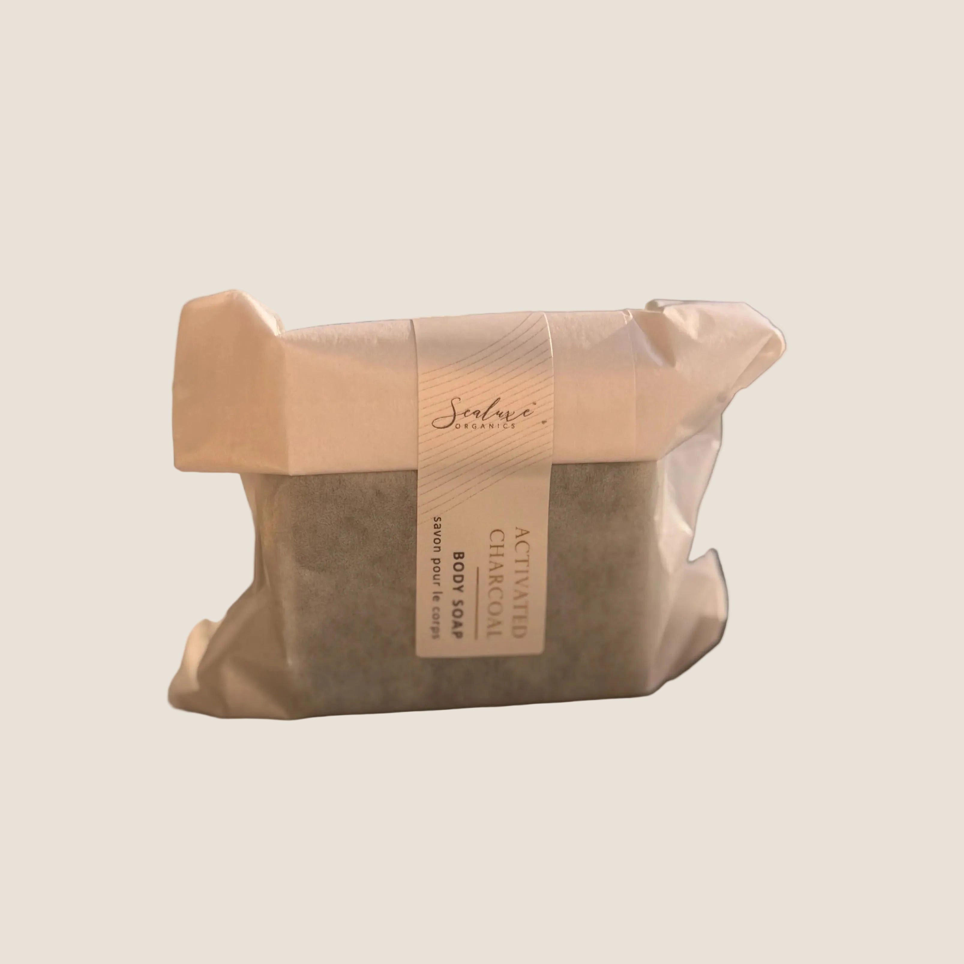 A wrapped rectangular body soap bar sits on a windowsill with a blurred outdoor background. The soap is covered in a light beige paper sleeve with a white paper band label that reads 'Serenade Soaps' and 'Activated Charcoal'.