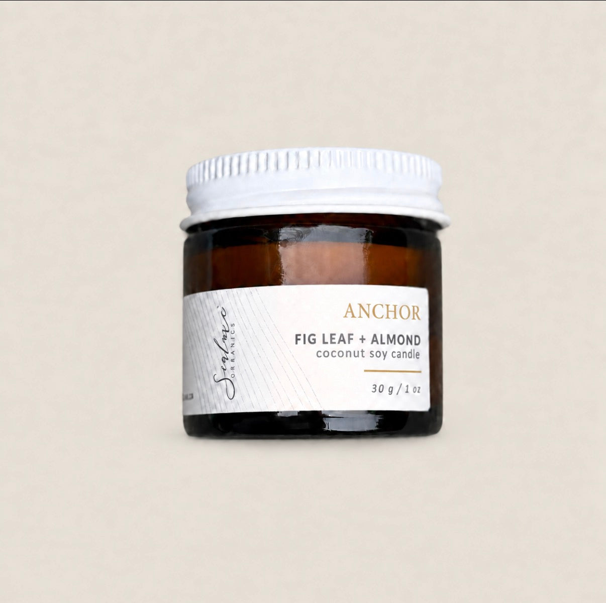 An amber glass jar of Anchor Fig Leaf + Almond coconut soy candle with a white ribbed metal lid and a white label, set against a solid beige background.