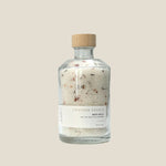 A clear glass jar with a wooden lid, labeled as 'Lavender Vanilla Bath Salt', containing white bath salts with visible small botanical particles.