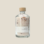 A clear glass bottle with a wooden cap containing rose bath salts. The label is minimalistic with the brand name 'ROSE' and the product name 'BATH SALTS' on it.