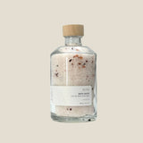 A clear glass bottle with a wooden cap containing rose bath salts. The label is minimalistic with the brand name 'ROSE' and the product name 'BATH SALTS' on it.