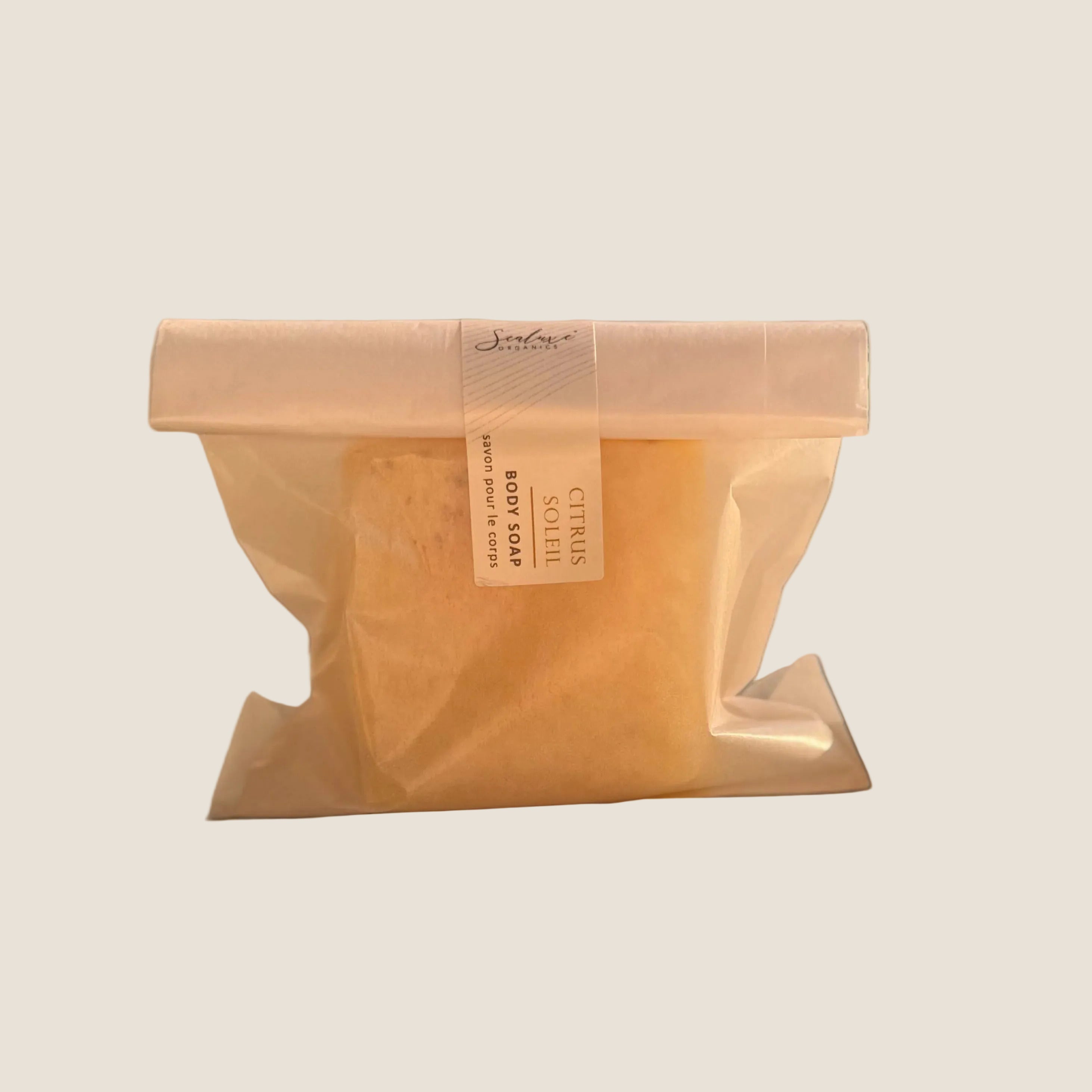 A single packaged body soap bar sits on a windowsill with warm natural light. The soap is wrapped in a translucent beige paper bag with a white paper band label that reads 'Jasmin' and 'Citrus'.