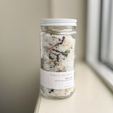 A glass jar with a metal lid labeled 'FOREST BATHING BATH TEA' containing white bath salts and natural botanicals.