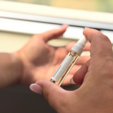 A close-up of a hand holding a small clear spray bottle labeled 'CLEAN HAND' with a white cap, while another hand is positioned behind it against a softly blurred light background.