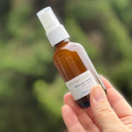 Hand holding a small brown bottle with a white spray nozzle against a blurred green background