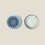 The image shows a glass jar with a metal lid, containing a natural lip balm. The lip balm is white and there is a snowflake design on the lid.