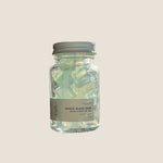 A small clear glass jar of Aqua Beach Glass Soap sits on a windowsill with natural light shining in from the right. The jar has a white label and a silver metal lid, and the background shows a softly blurred outdoor scene through the window.