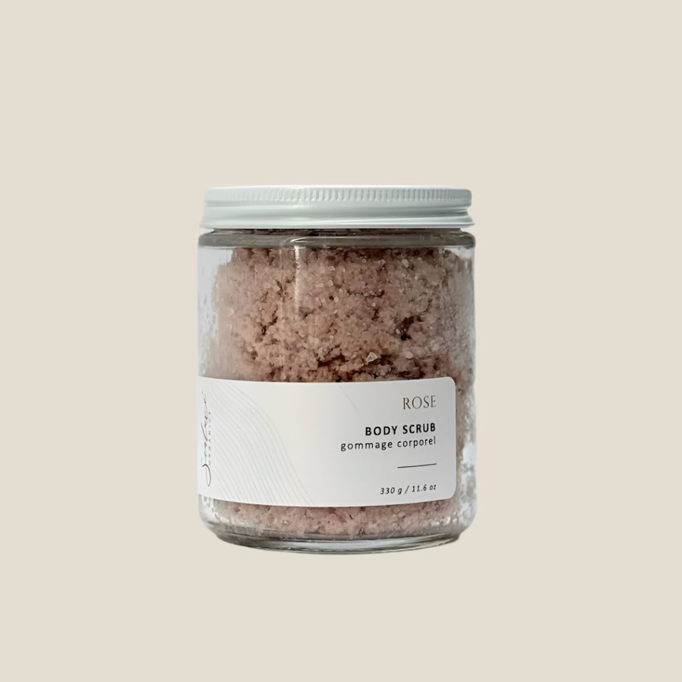 A clear glass jar with a white label and a black cap containing a pale pink and black scrub with the words "Ritual Rose" written on the label. The background is plain white.