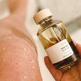 A hand holds a glass bottle of rose-scented body oil, infused with delicate petals and seaweed, shimmering in the golden light of the beach. The ocean waves roll gently in the background, evoking a sense of tranquility and self-care with every drop of this luxurious, hydrating blend.