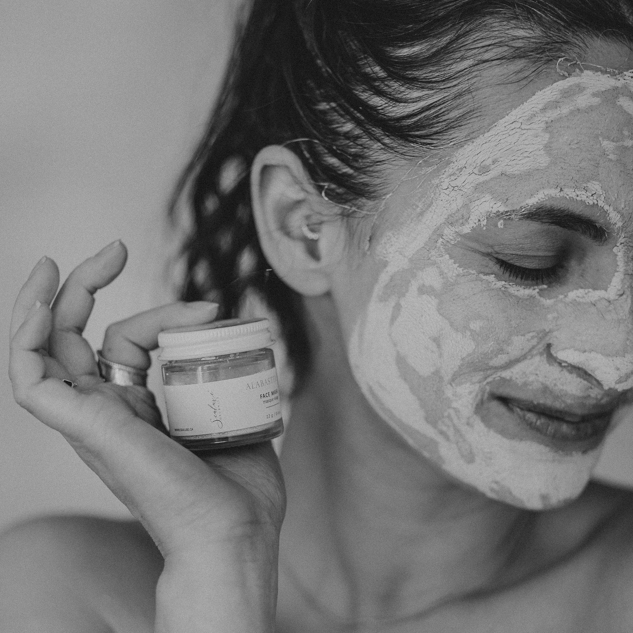 Black and white close-up of woman with Sealuxe Alabaster clay face mask applied to her face, holding the product jar — natural face mask in use