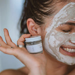 Smiling woman wearing Sealuxe Alabaster Face Mask while holding the jar, waving hand toward camera