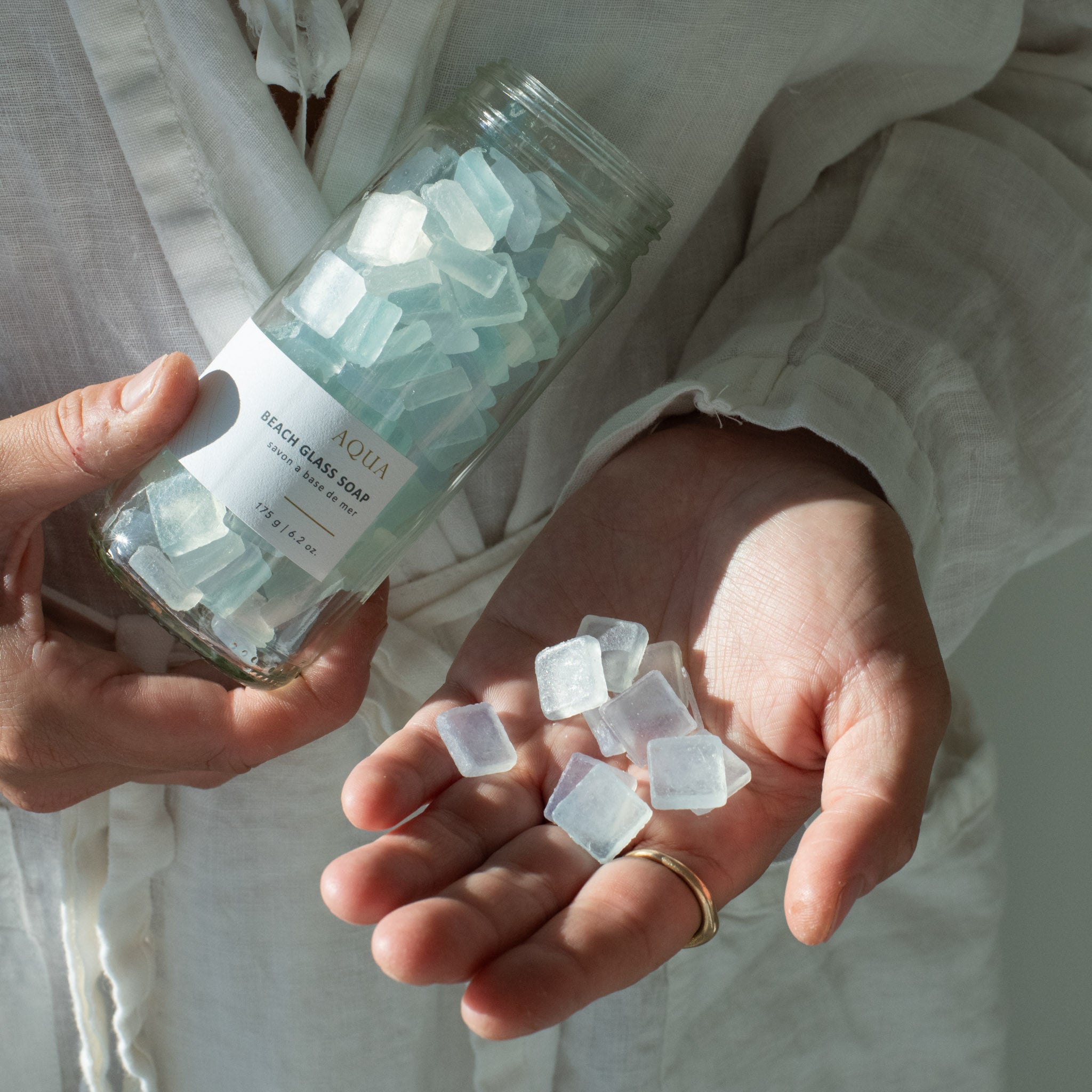Hand holding open jar of Sealuxe Aqua Beach Glass Soap with turquoise sea glass soap pieces