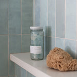 Sealuxe Aqua Beach Glass Soap jar on white tiled shower shelf beside a natural sea sponge