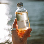 Sealuxe Driftwood Body Oil 125ml glass bottle resting on sunlit driftwood near water