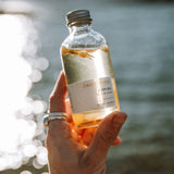 Sealuxe Driftwood Body Oil 125ml glass bottle resting on sunlit driftwood near water