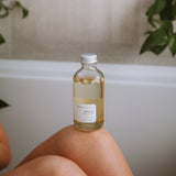 Sealuxe Driftwood Body Oil bottle resting on a bath tray beside a drawn bath