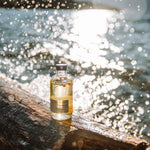 Small Sealuxe Driftwood Body Oil glass bottle resting on a driftwood log with sparkling sunlit ocean in the background — natural body oil coastal lifestyle