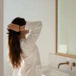 Woman in white linen styling her hair in a bright bathroom — Sealuxe lifestyle