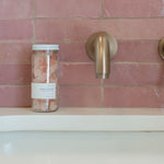 Sealuxe Rose Beach Glass Soap jar with pink sea glass soap pieces on a pink tiled bath ledge beside a gold faucet