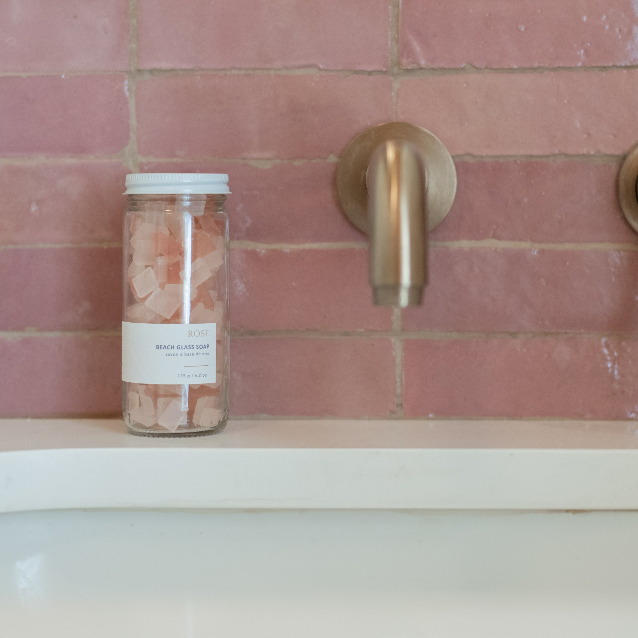 Sealuxe Rose Beach Glass Soap jar with pink sea glass soap pieces on a pink tiled bath ledge beside a gold faucet