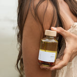Woman holding a glass bottle of Sealuxe Rose Body Oil beside her bare shoulder — natural rose body oil