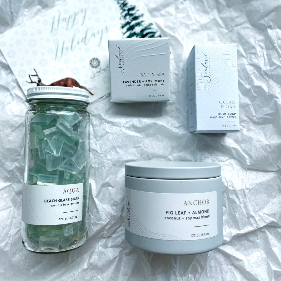 Flat lay of Sealuxe 'Sand and Snow' winter beach gift set, featuring Aqua Beach Glass soap, Ocean Flora soap bar, Westcoast White candle, and Salty Sea bath bomb, artistically arranged to evoke a coastal winter escape.