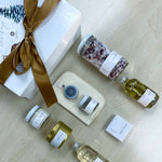 Sealuxe Spa at Home gift box featuring bubbles, body oil, bath tea, bath bomb, Driftwood butter, seaweed face mask, cloth, Gold Anchor candle, and lip balm, arranged for a luxurious home spa experience.