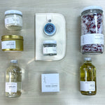 Flat lay view of Sealuxe Spa at Home gift box products including bubbles, body oil, bath tea, bath bomb, Driftwood butter, seaweed face mask, cloth, Gold Anchor candle, and lip balm, artistically displayed for a full spa experience.