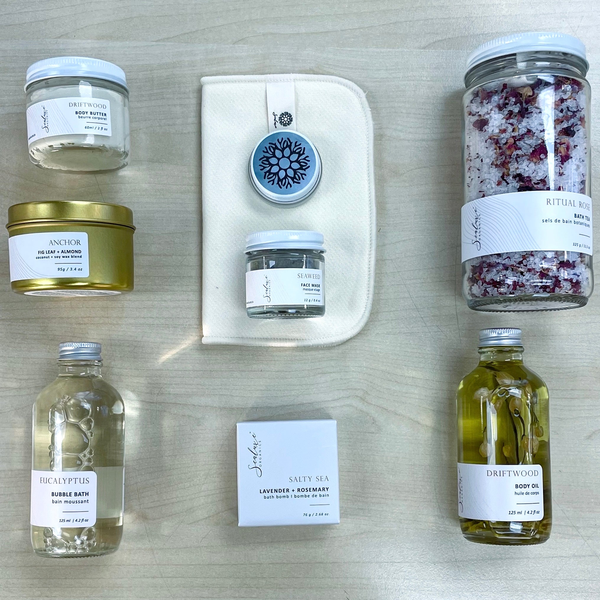 Flat lay view of Sealuxe Spa at Home gift box products including bubbles, body oil, bath tea, bath bomb, Driftwood butter, seaweed face mask, cloth, Gold Anchor candle, and lip balm, artistically displayed for a full spa experience.