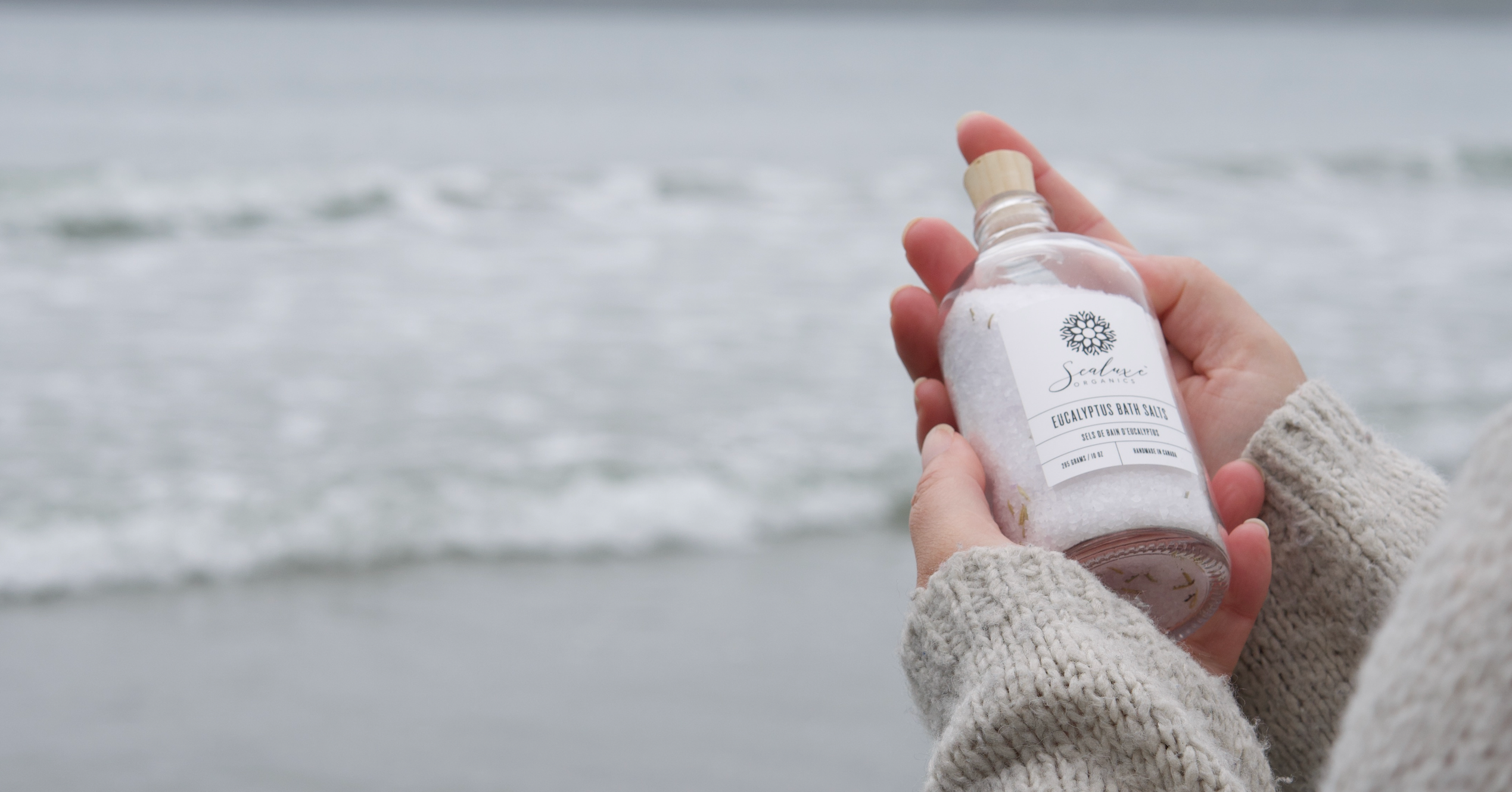holding a glass bottle of sealuxe eucalyptus bath salts with the ocean in the background