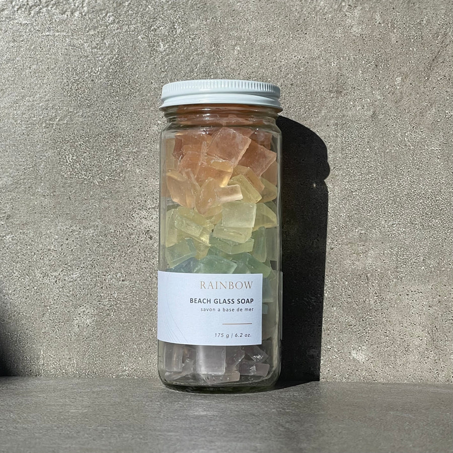 A glass jar with a lid is filled with small, irregularly shaped pieces of Sealuxe Rainbow beach glass soap. The soap pieces come in bright colors of the rainbow and are arranged in a gradient, with red at the top, followed by orange, yellow, green, blue, and purple at the bottom. The pieces are small and chunky, with a textured surface. A label on the front of the jar reads 'Sealuxe Rainbow beach glass soap' and features ocean imagery and the Sealuxe logo.